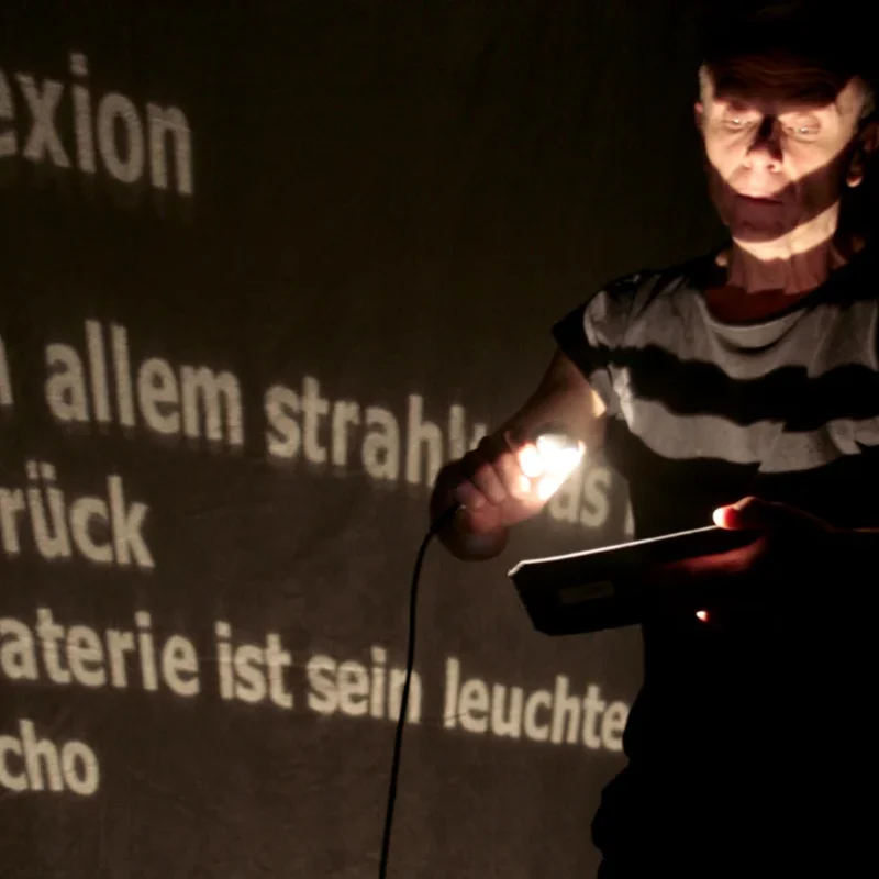 Performer hält Taschenlampe vor projiziertem Text auf dunkler Bühne – Lichtperformance „17 Sätze über das Licht", Theater der Schatten Bamberg